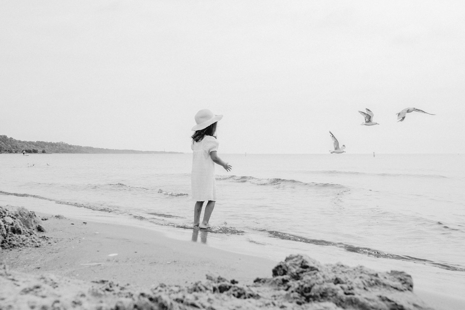 A black and white photograph of someone standing at the beach watching seagulls fly over the ocean.