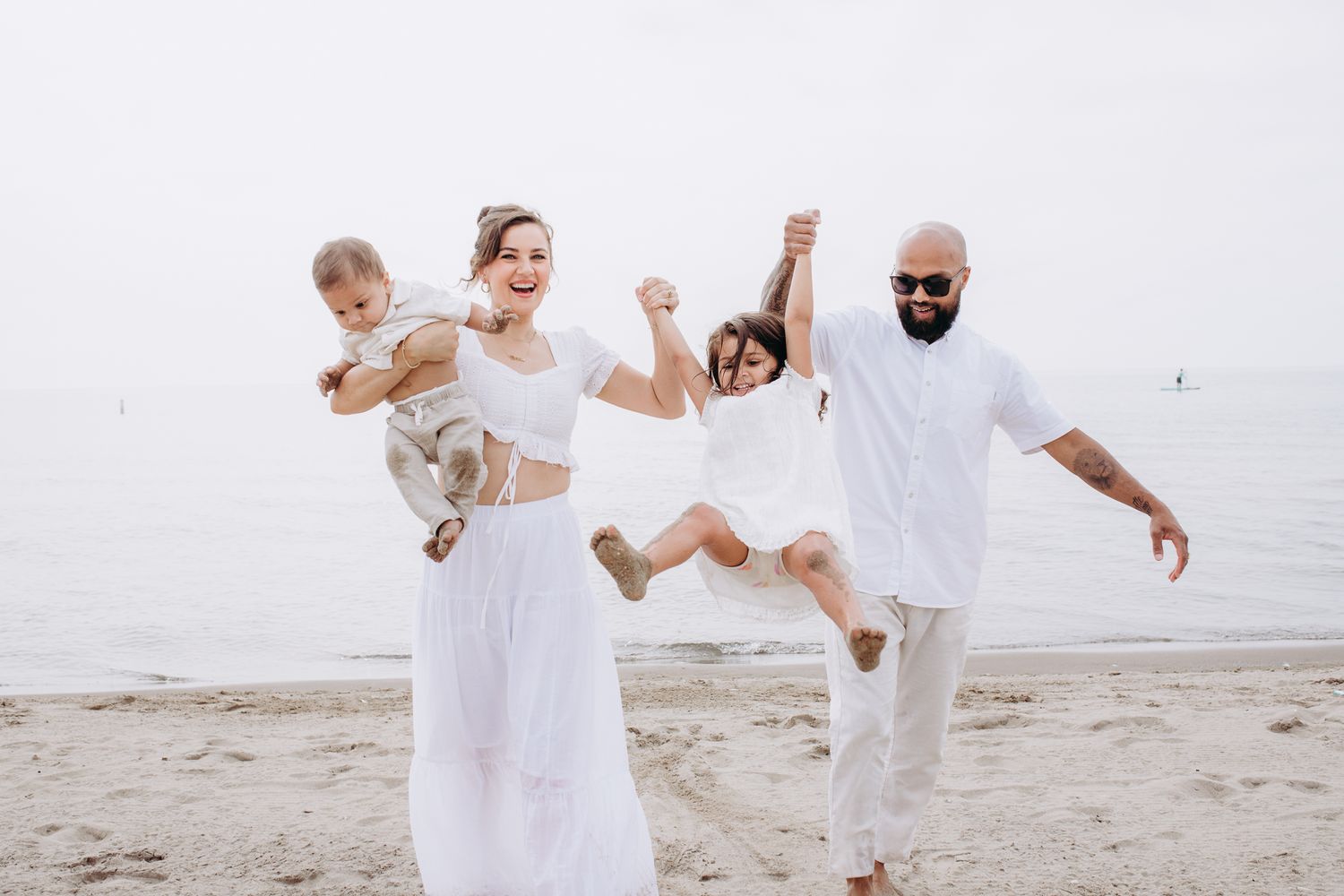 A joyful family wearing all white outfits plays together and jumps on a sandy beach by the water.