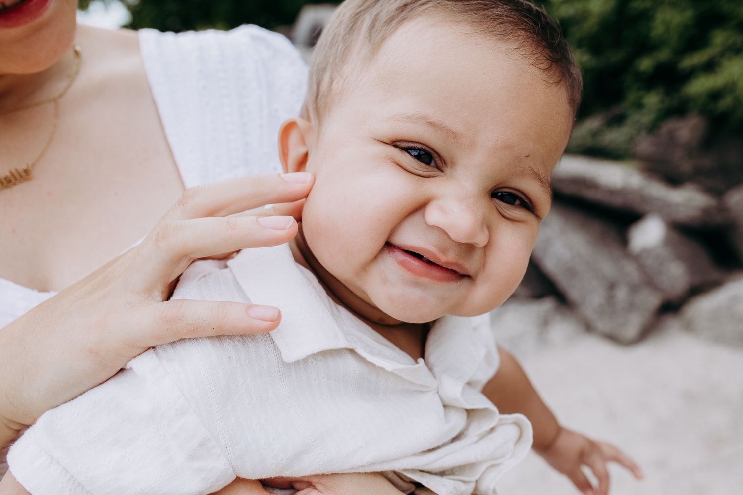 Baby wearing white clothing smiles joyfully while being held by an adult.