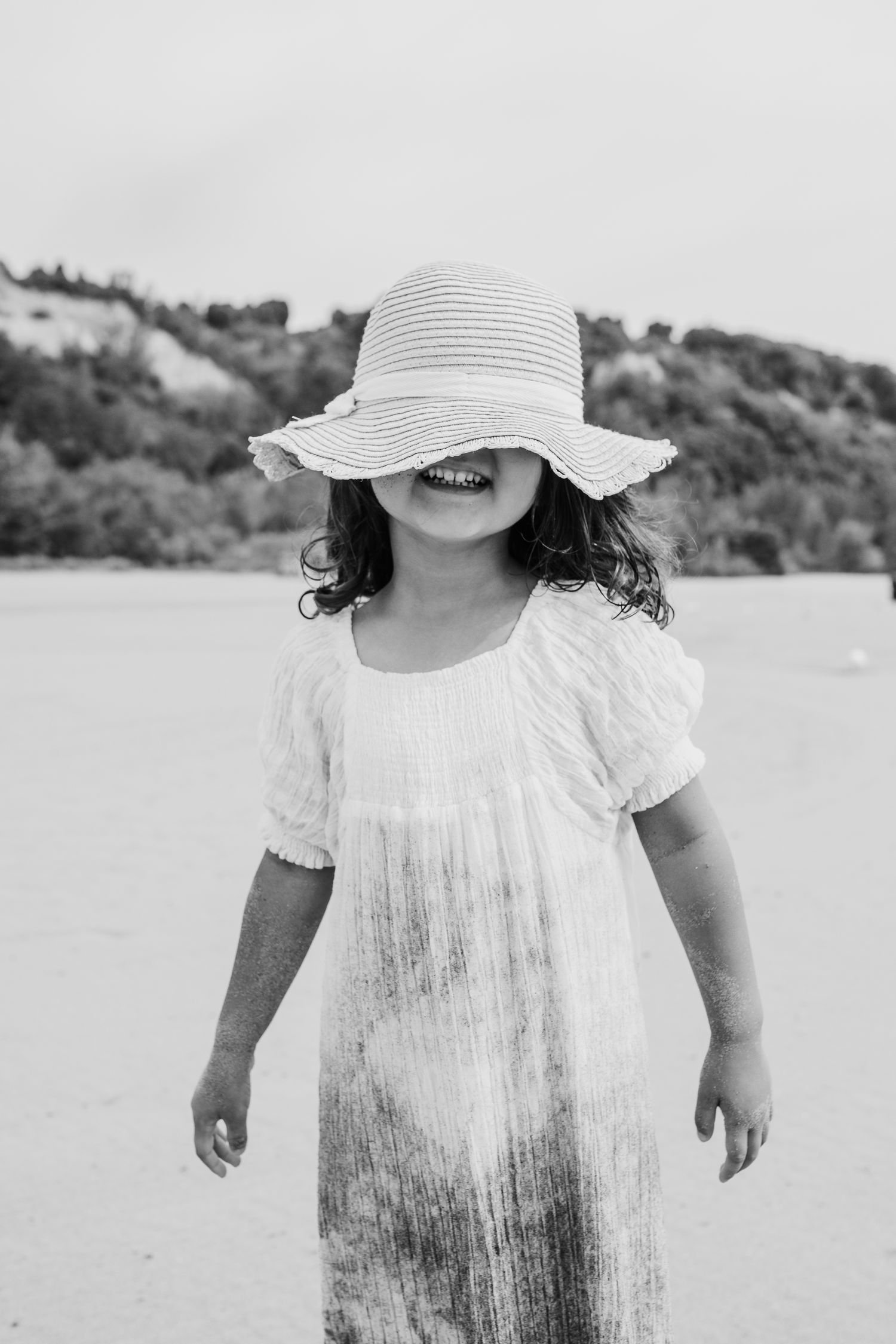 Black and white photo of child in white dress and sun hat at beach.