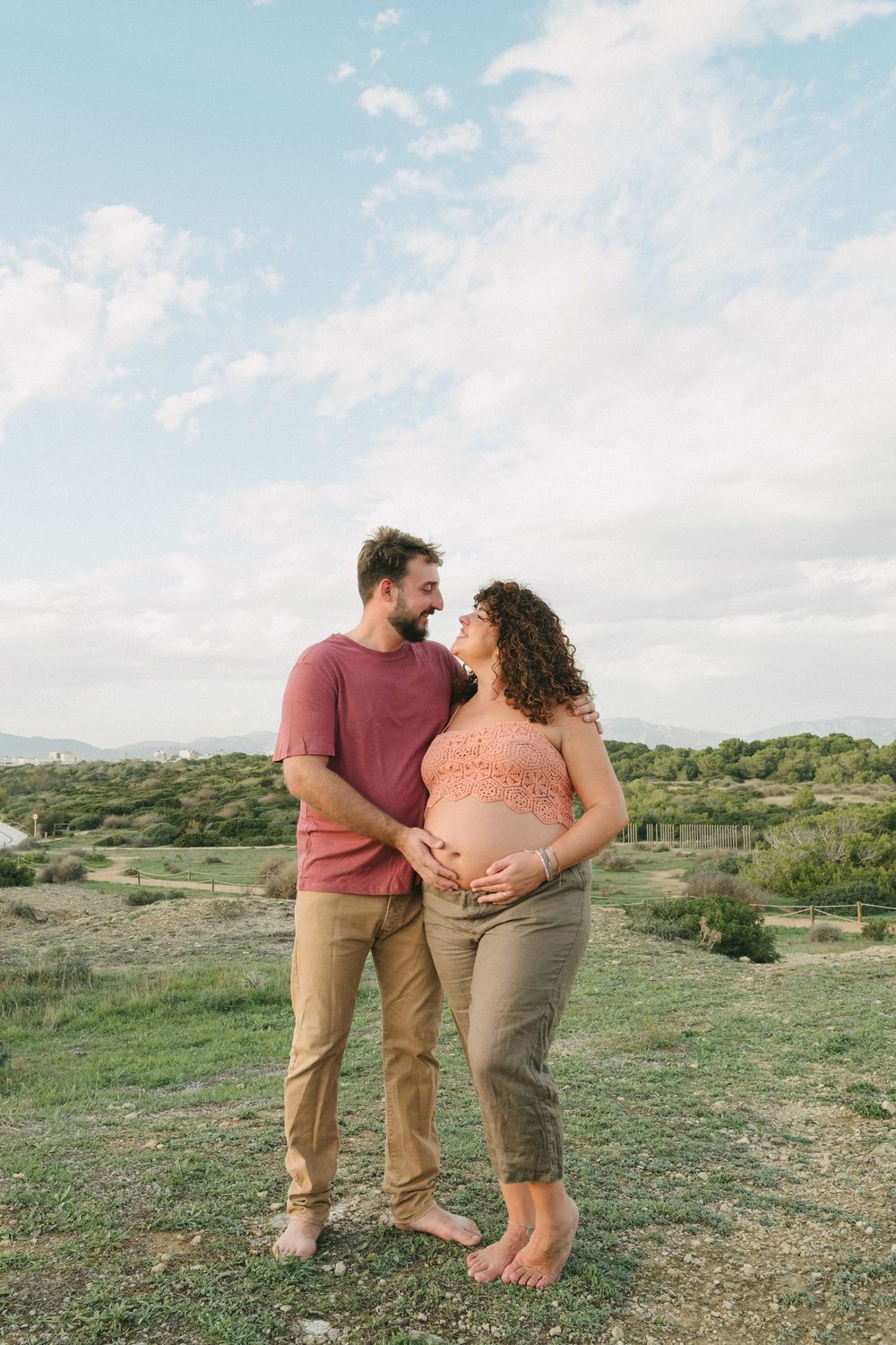 Expecting couple embraces on grassy hilltop with scenic landscape and blue sky in background during maternity photoshoot.