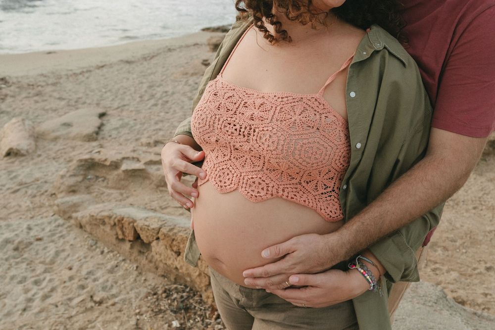 A pregnant belly in a pink lace top is embraced on a sandy beach while wearing an olive green jacket.