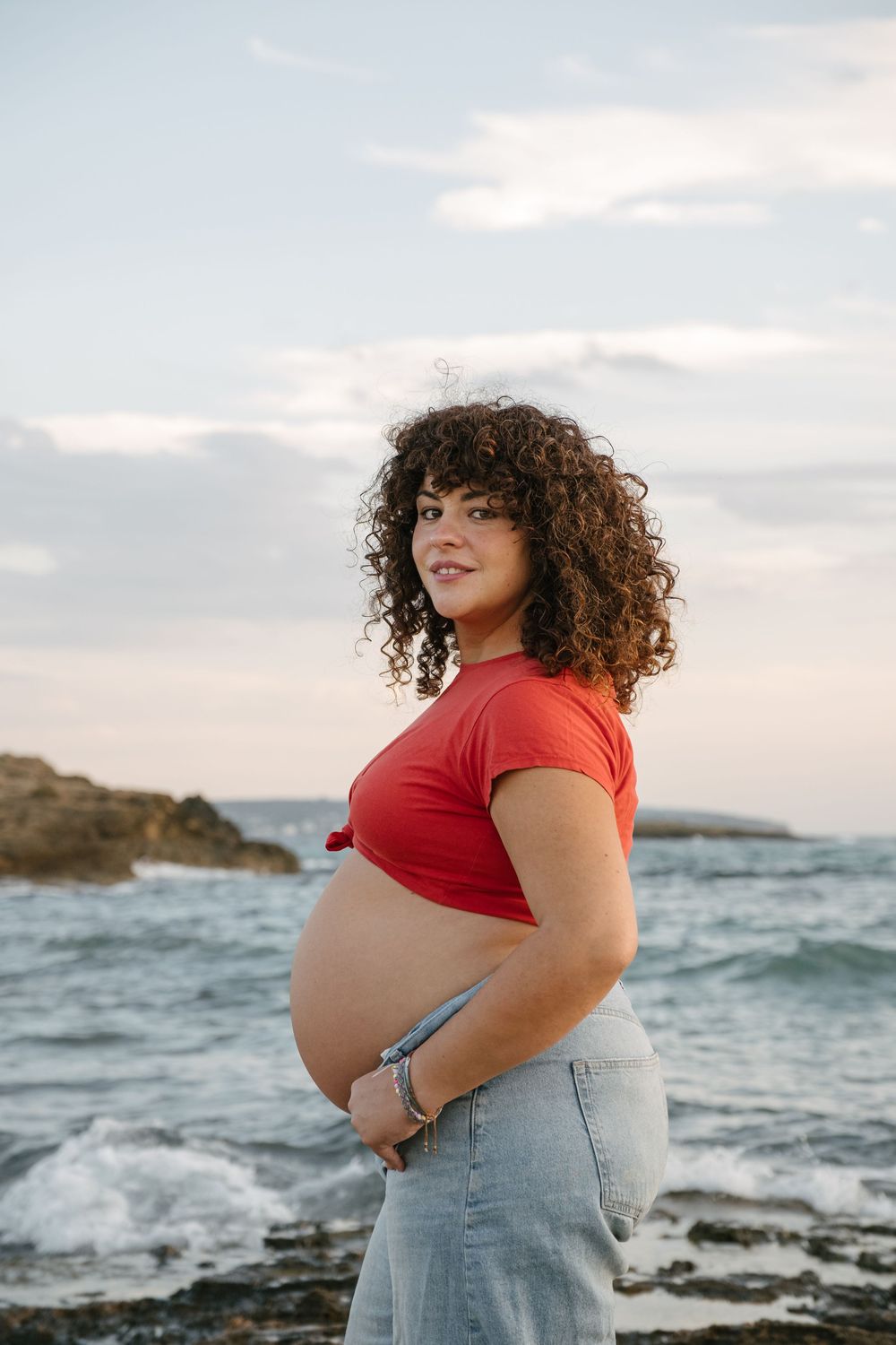 A scenic maternity photoshoot at a rocky beach coastline during sunset with waves crashing in the background.