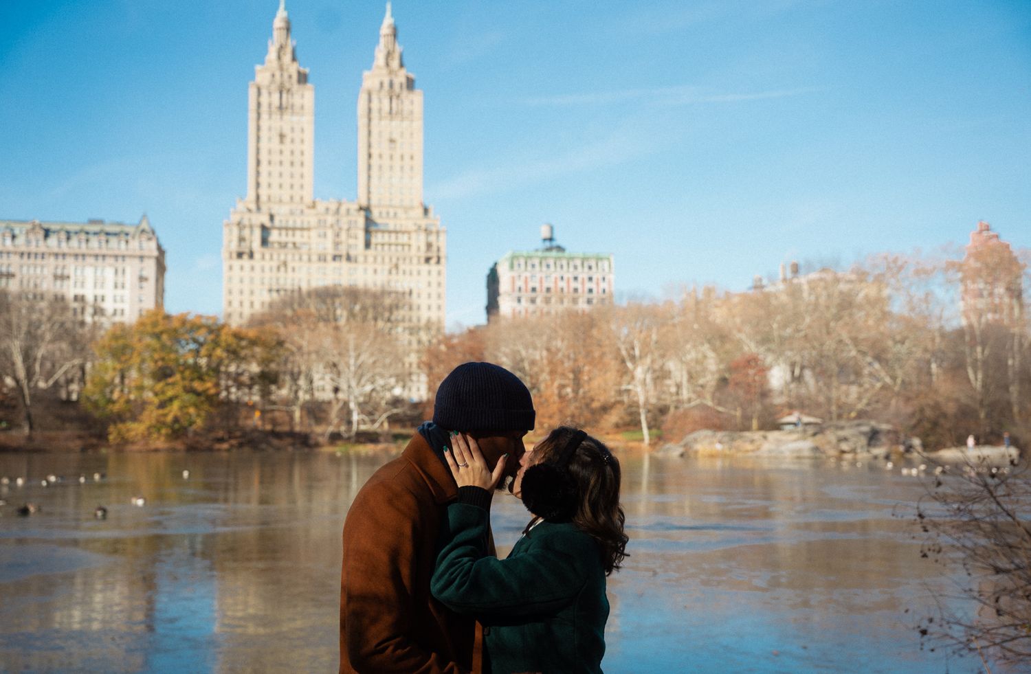 A couple shares a romantic moment near a lake with the iconic Central Park skyline in winter.