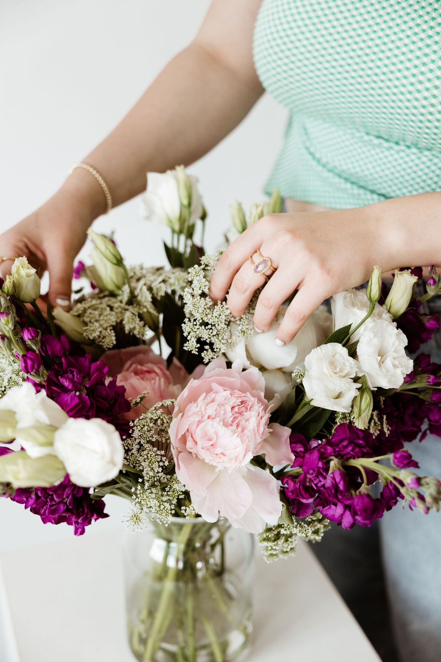 Close-up of hands with gold rings arranging a bouquet of purple, pink, and white flowers in a vase.