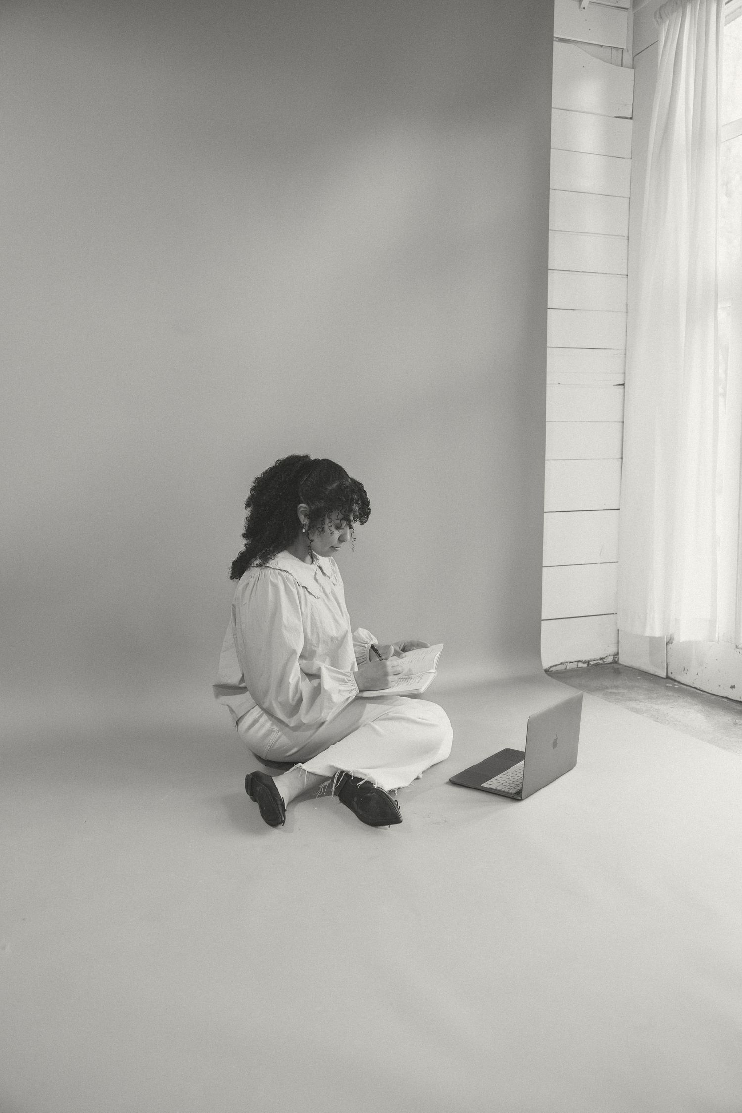 Black and white photo of a woman sitting cross-legged on the floor writing in a notebook with a laptop nearby.