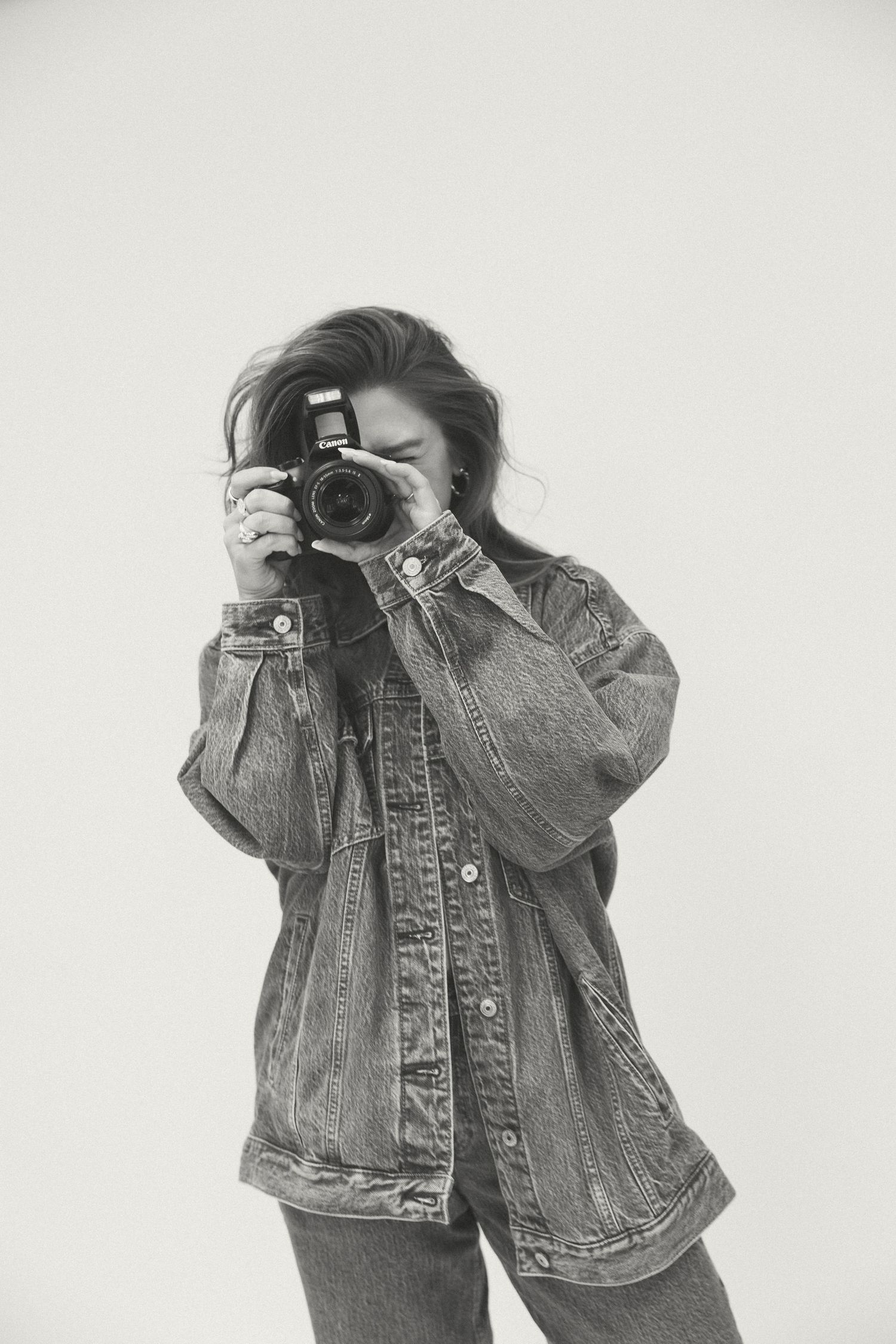 Black and white photo of woman in oversized denim jacket photographing with DSLR camera.