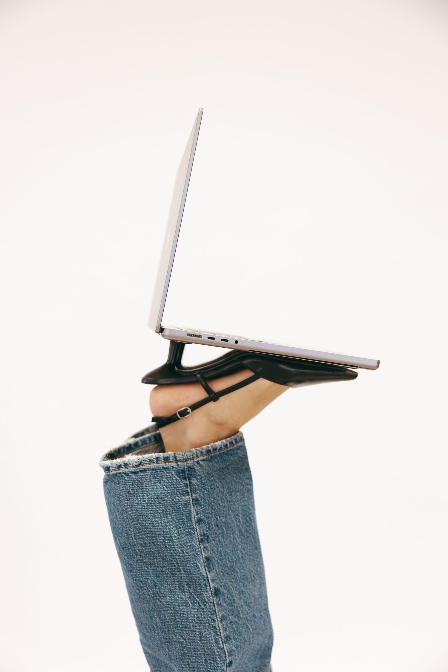 Person in jeans balancing open silver laptop on raised foot against white background.