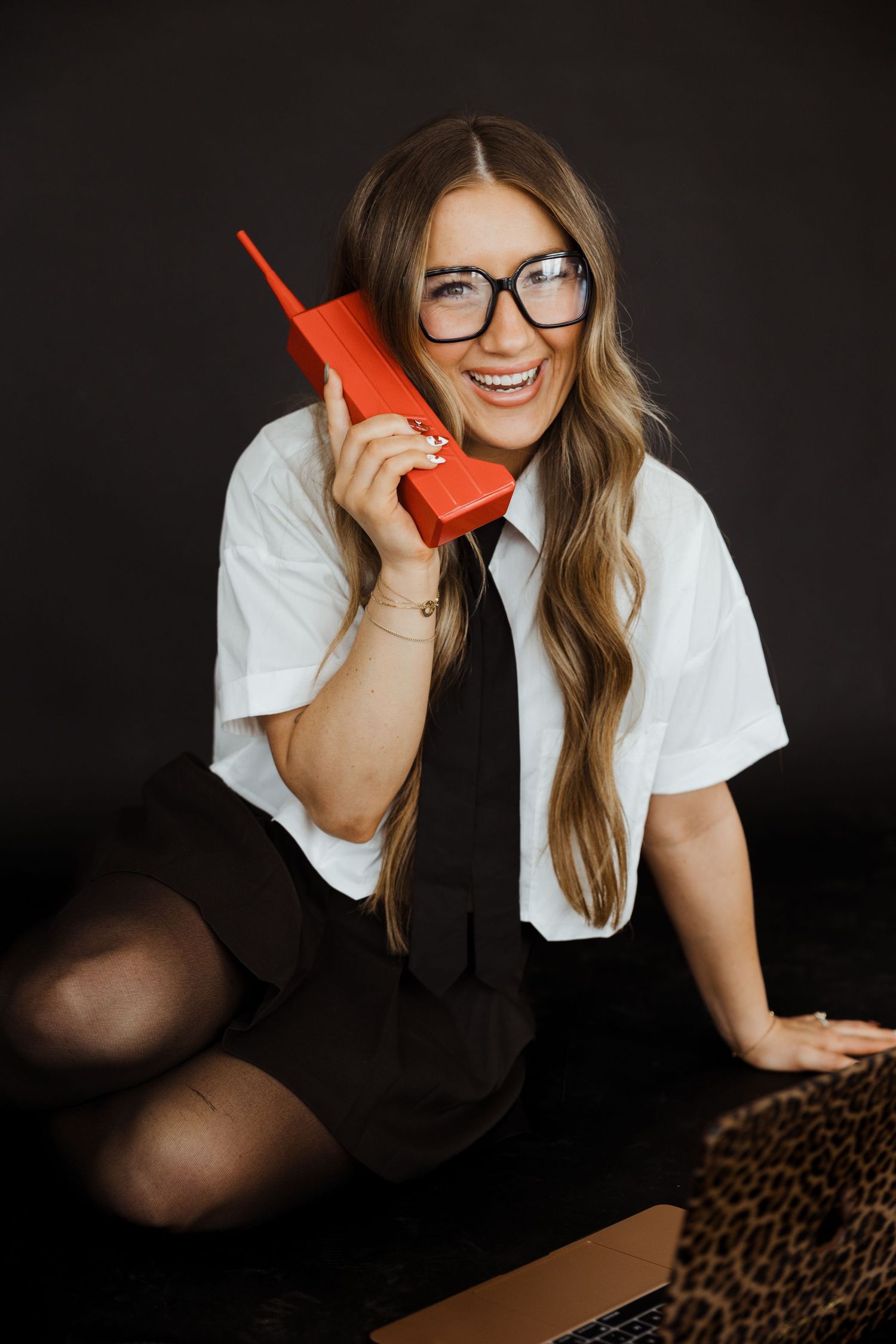 Woman in white shirt and glasses holding red retro phone while sitting with laptop.