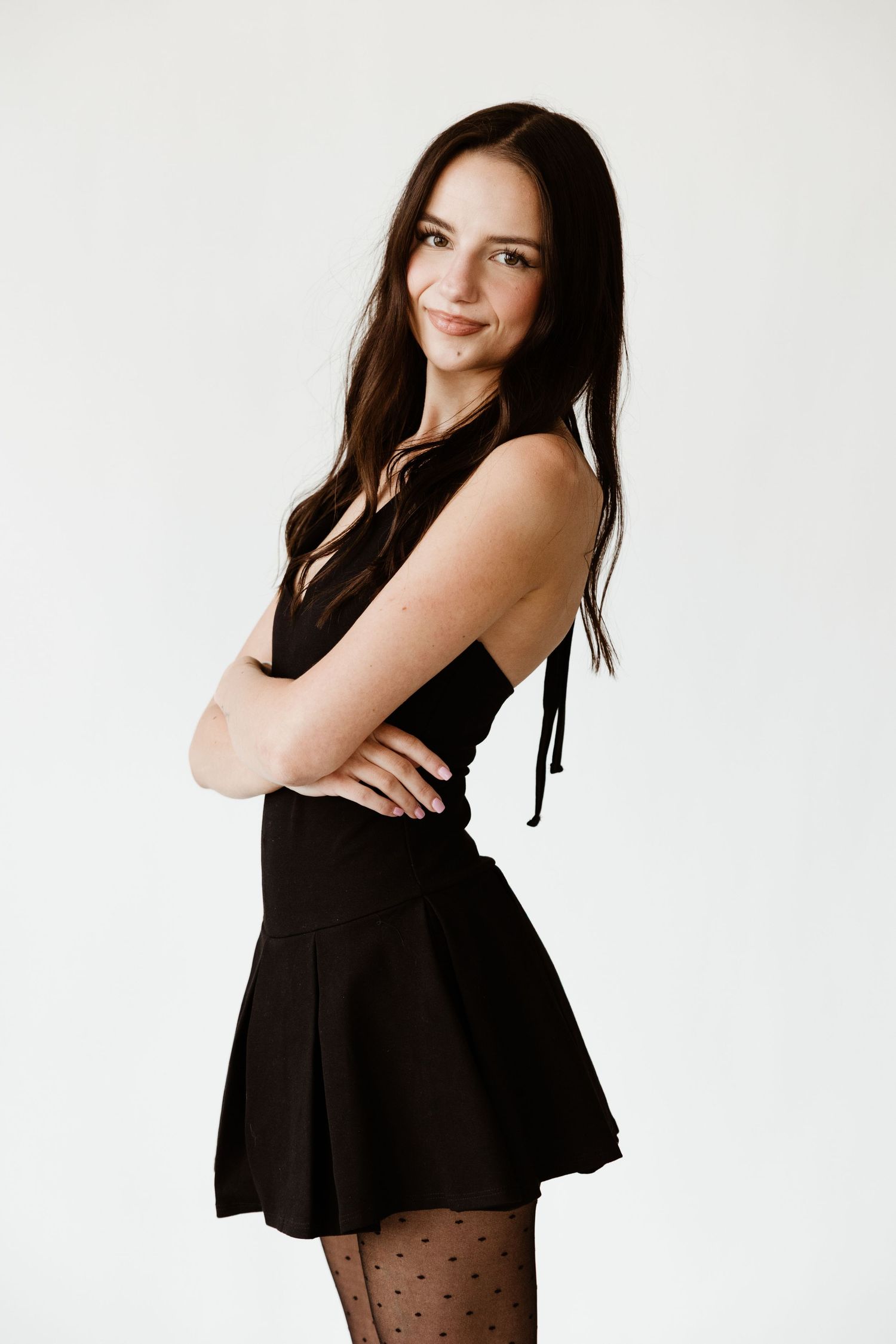 A young brunette woman in a black mini dress and polka dot tights posing with arms crossed.