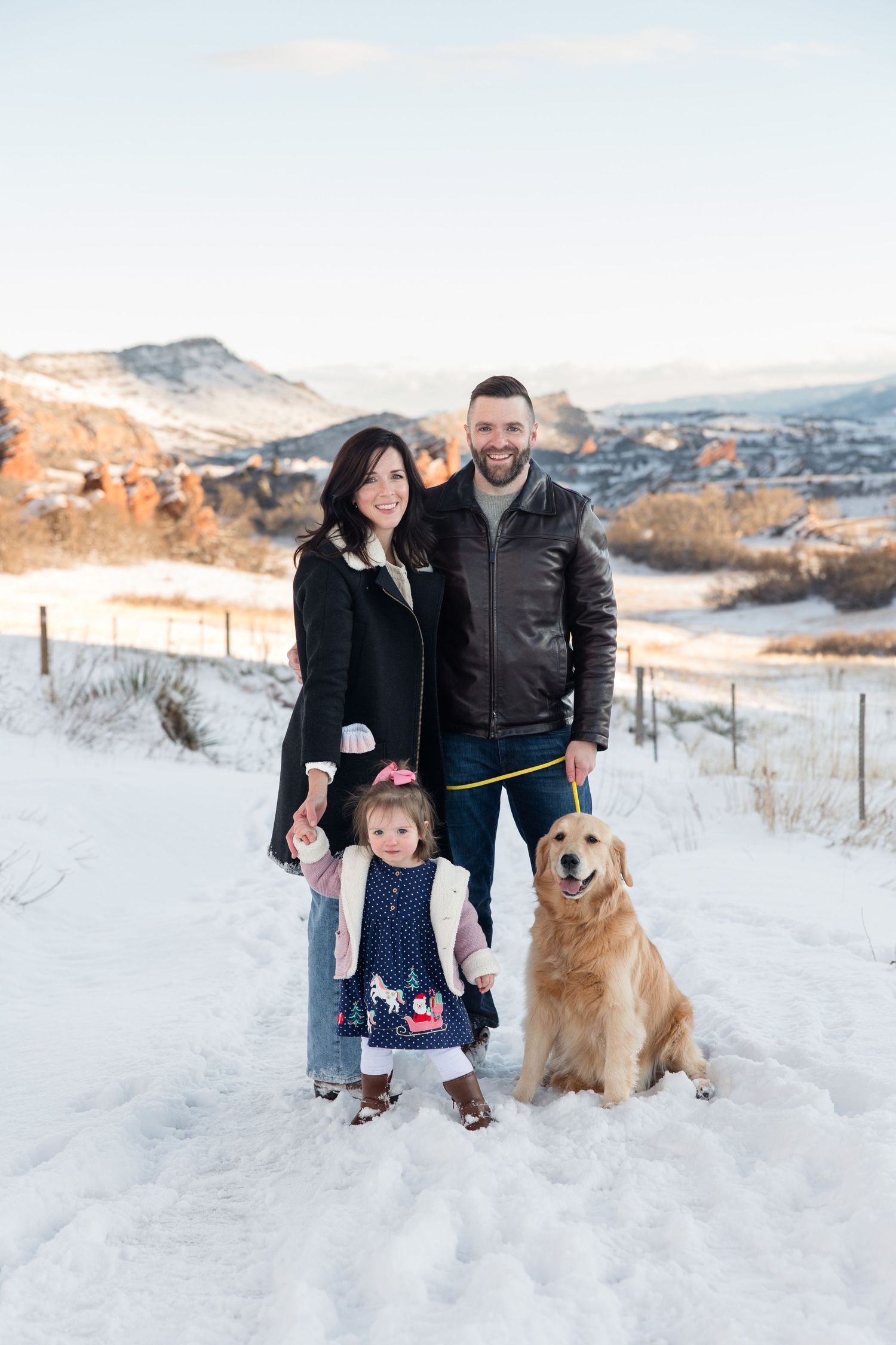 A couple and their golden retriever pose together in a snowy winter landscape with mountains in the background.