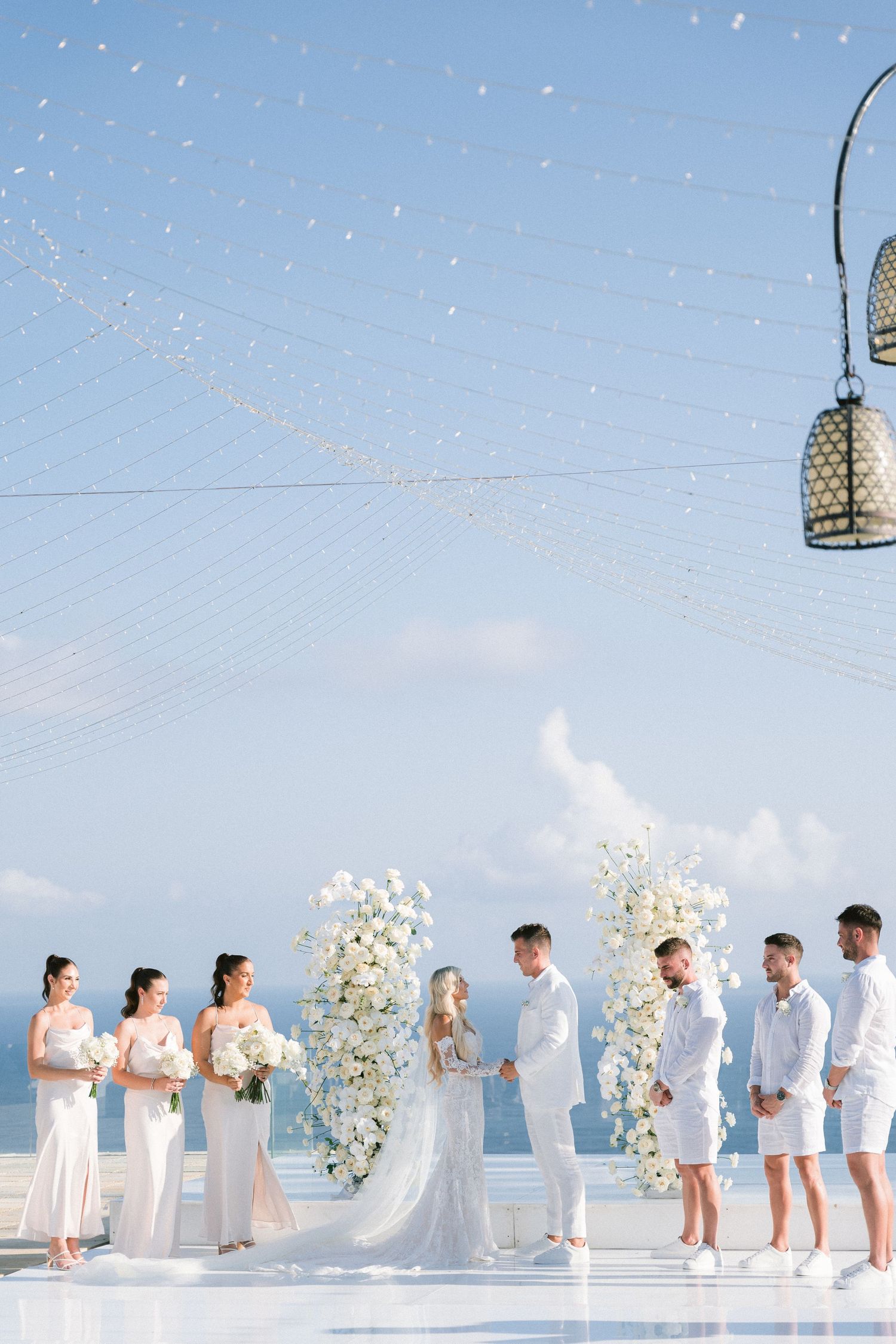 Elegant outdoor wedding ceremony with white floral arch and decor against blue sky and ocean backdrop.