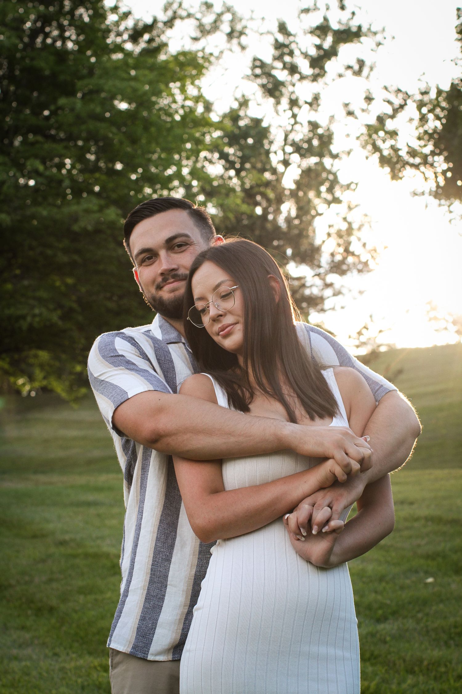 A woman with closed eyes and soft smile leans backward into her fiance, who is hugging her from behind and smiling at the camera.