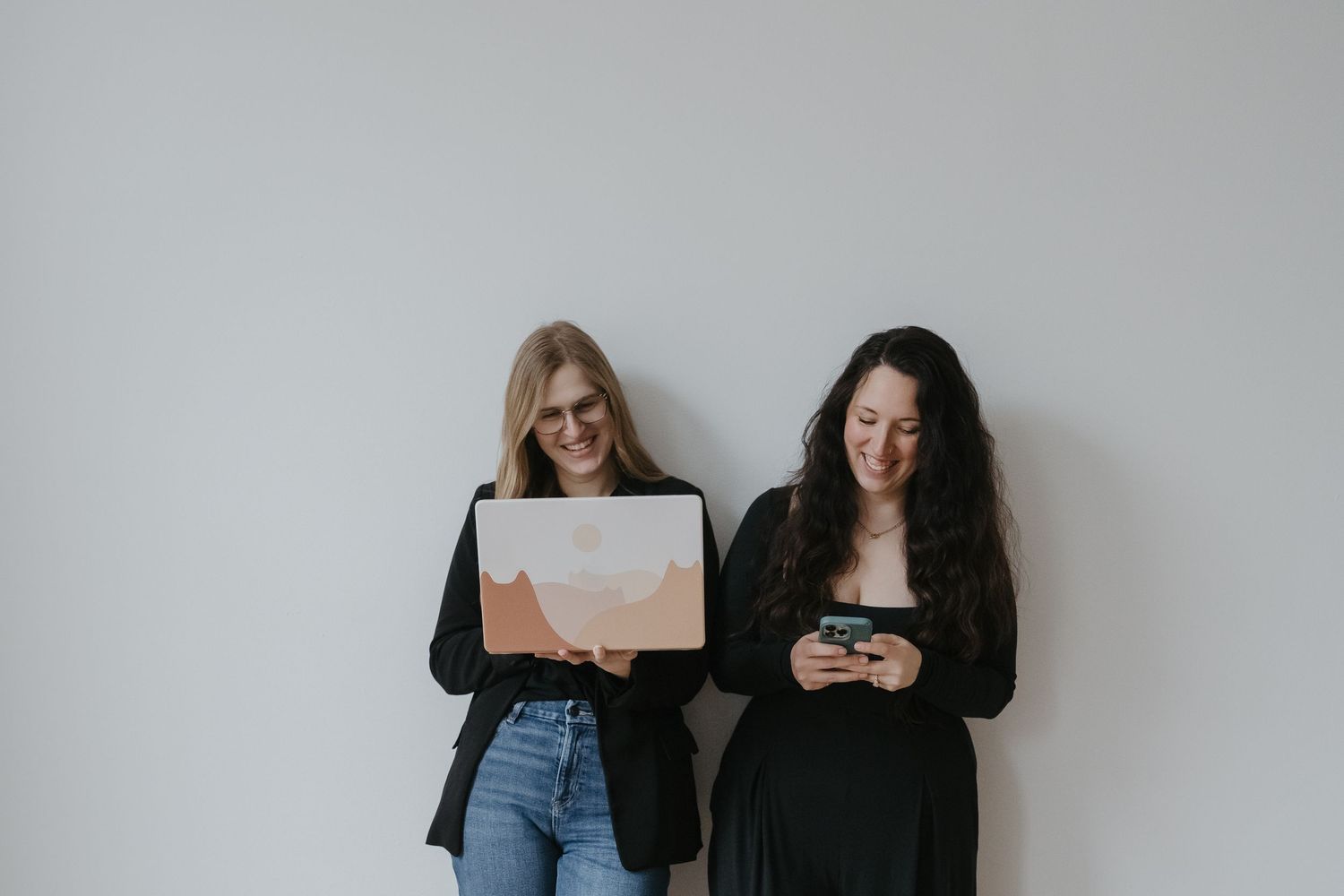 Two people in casual clothing standing against a white wall while looking at their mobile phones.