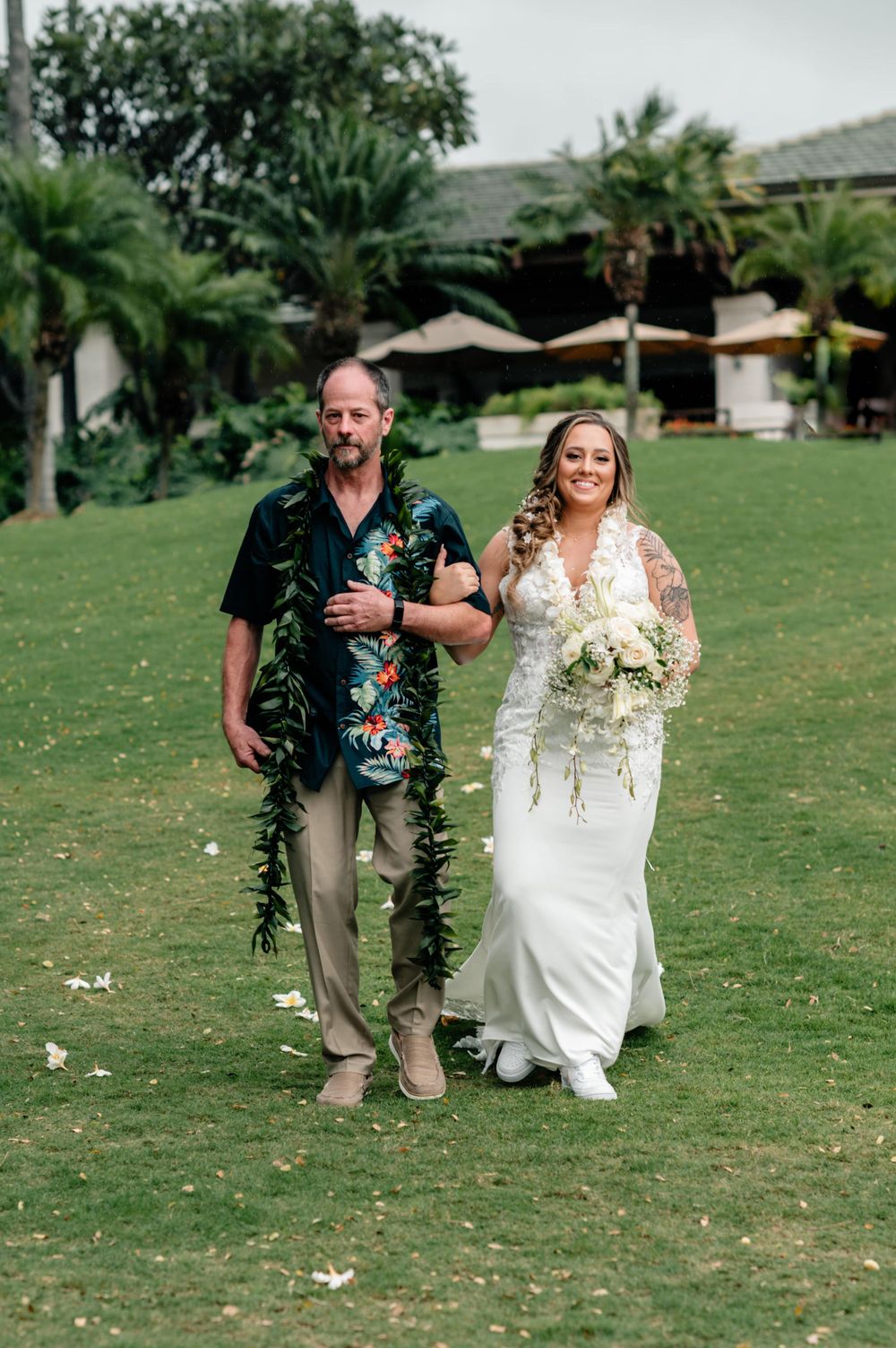 Wedding ceremony procession walking across green lawn with tropical foliage.