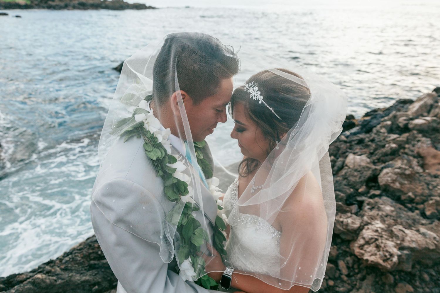Romantic oceanside moment with couple wearing traditional wedding attire and Hawaiian leis.
