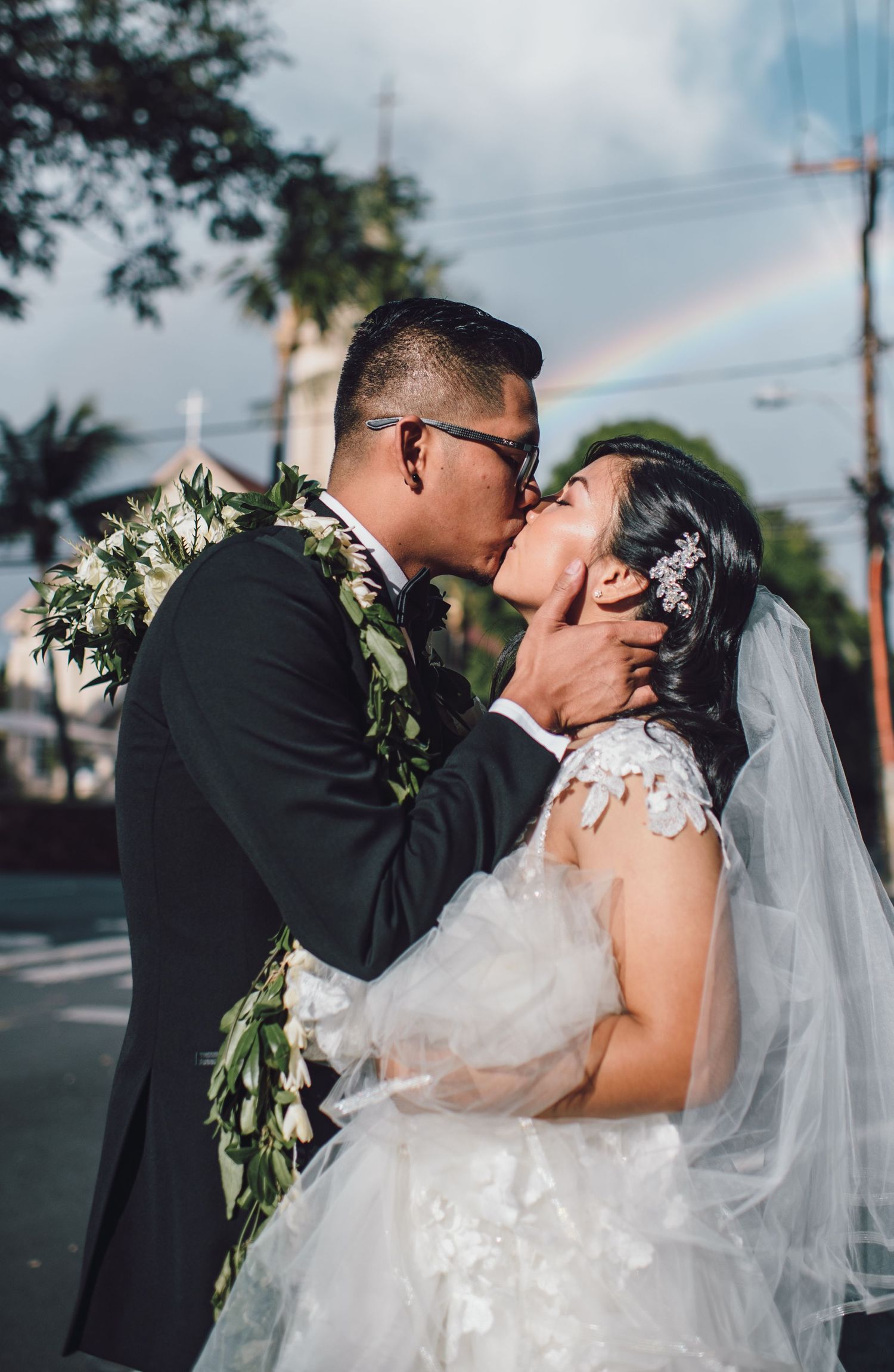 Wedding couple sharing intimate kiss with rainbow in background and traditional Hawaiian leis.