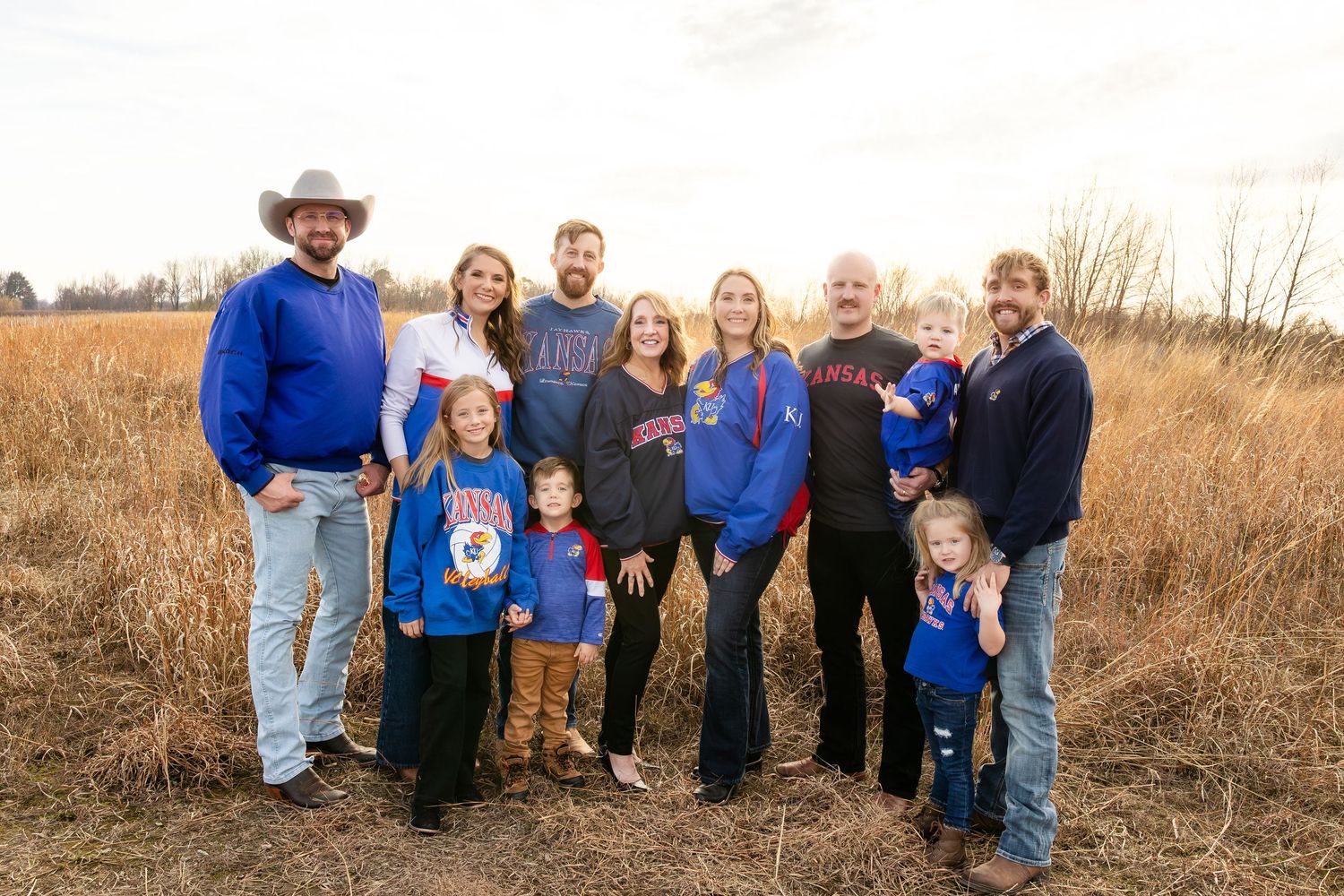 Family group poses together outdoors in a field during autumn wearing casual clothing in blue and neutral tones.