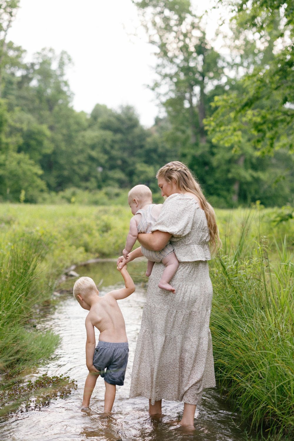 A parent and children playing together in a shallow stream on a warm summer day surrounded by green foliage.