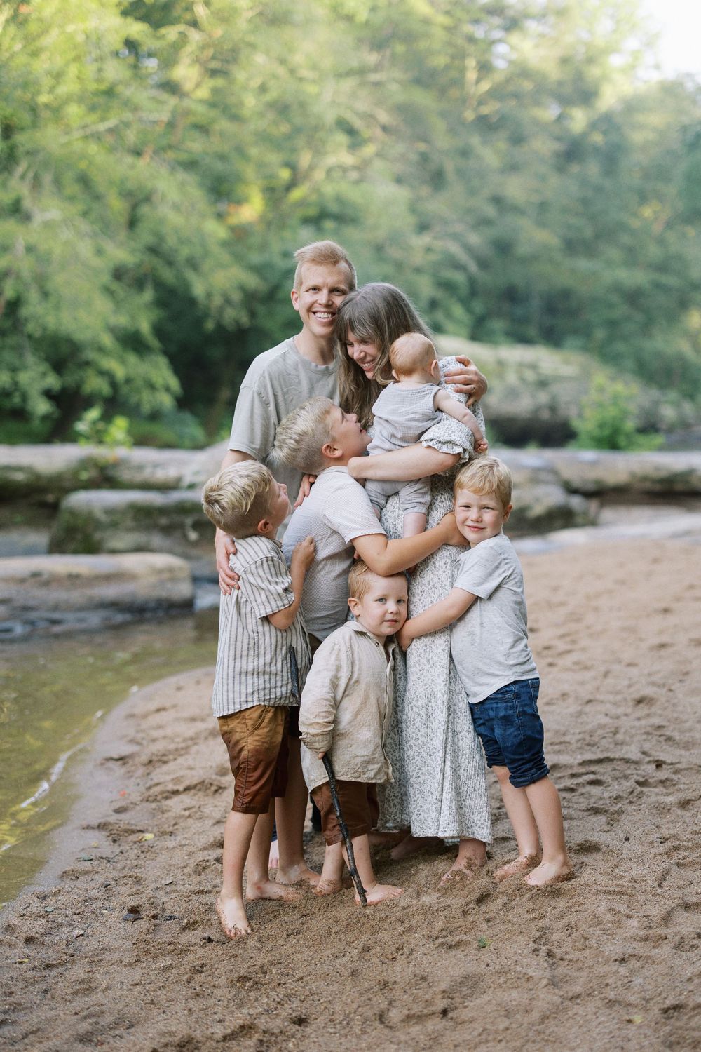 A family shares tender moments together on a sandy riverbank surrounded by lush greenery.