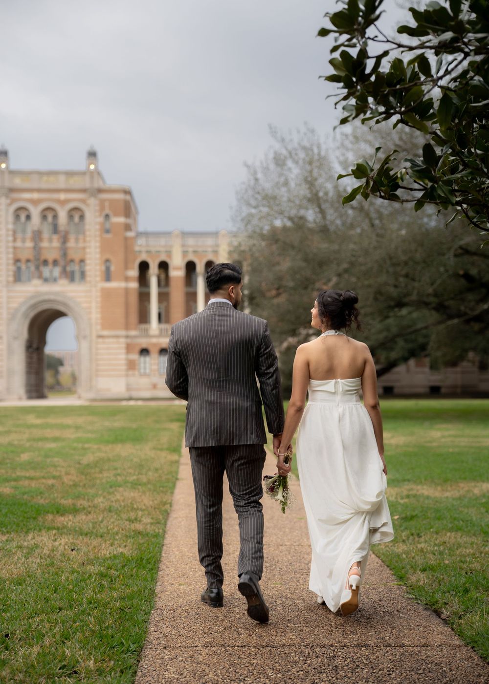 Rubi and Joe walking hand in hand on the paths of Rice University for their wedding portrait session, with sunlight filtering through trees