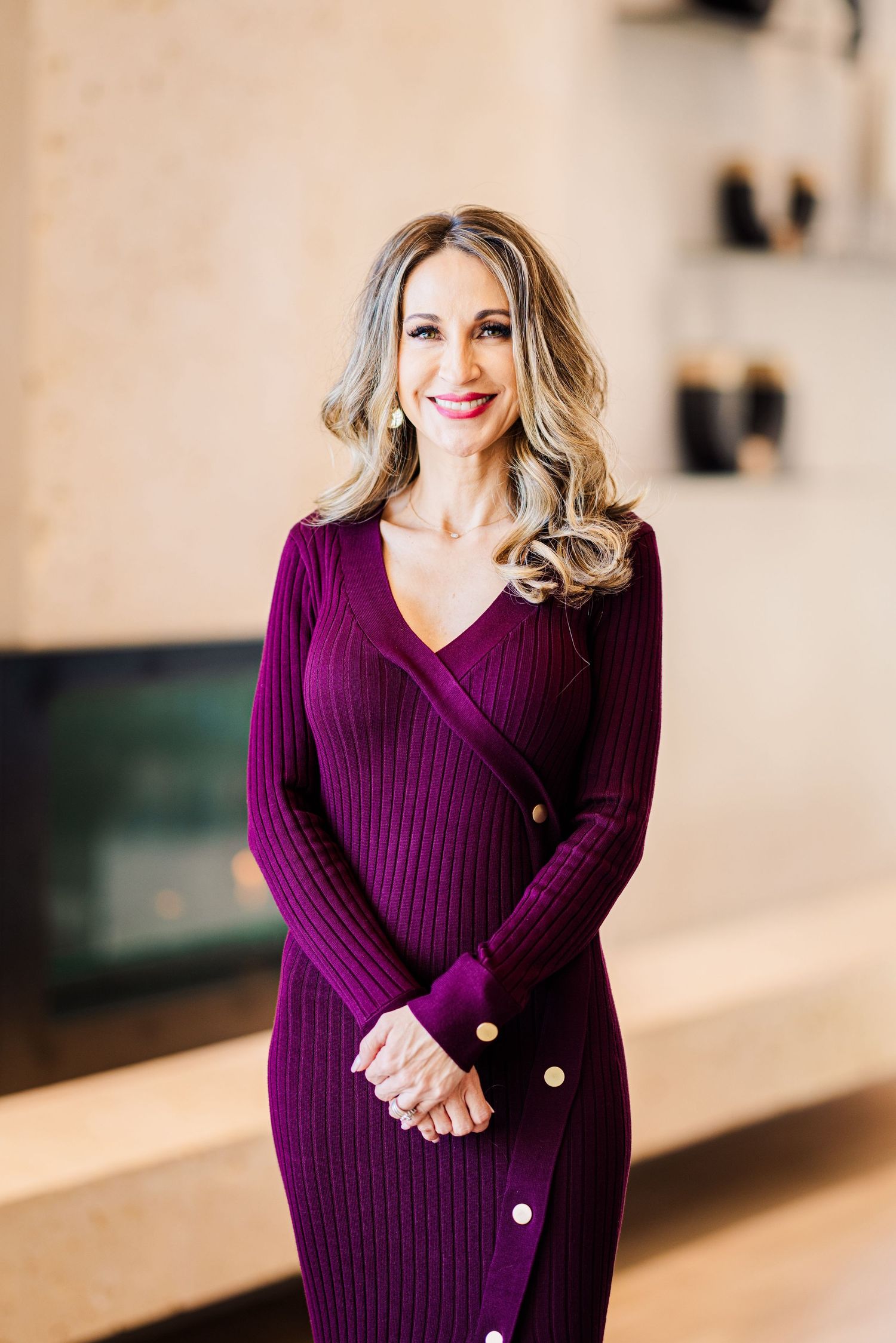 A smiling woman in a purple ribbed wrap dress poses confidently indoors with soft background lighting.