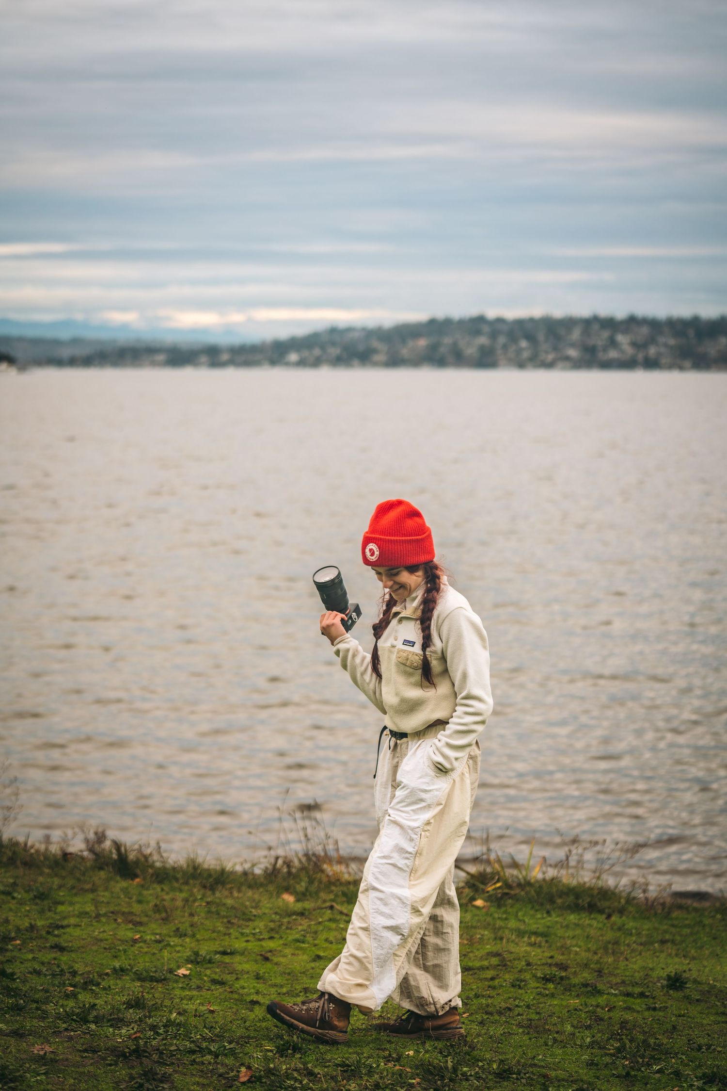 A woman in white pants and fleece with a red beanie holds a camera lens near a gray lake shore.