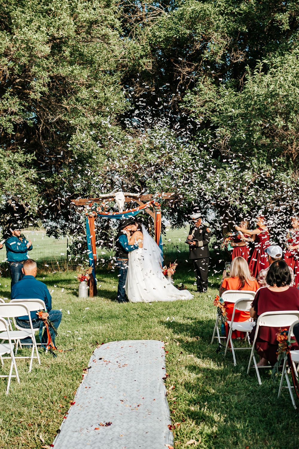 Guests throw confetti during an outdoor wedding ceremony with white folding chairs arranged on a grassy lawn.