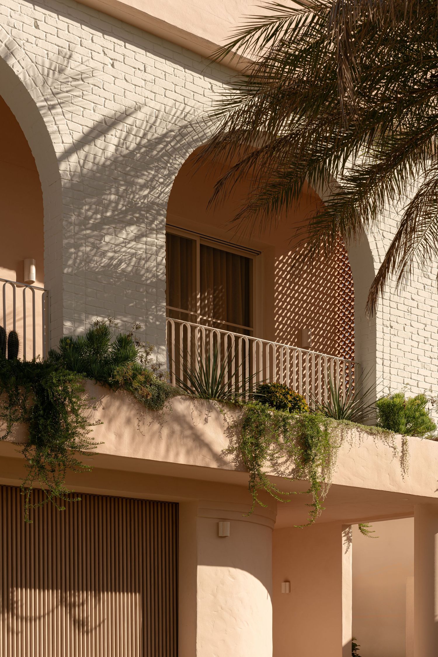 Modern architectural detail showing white brick walls with arched windows and palm fronds casting shadows in warm sunlight.