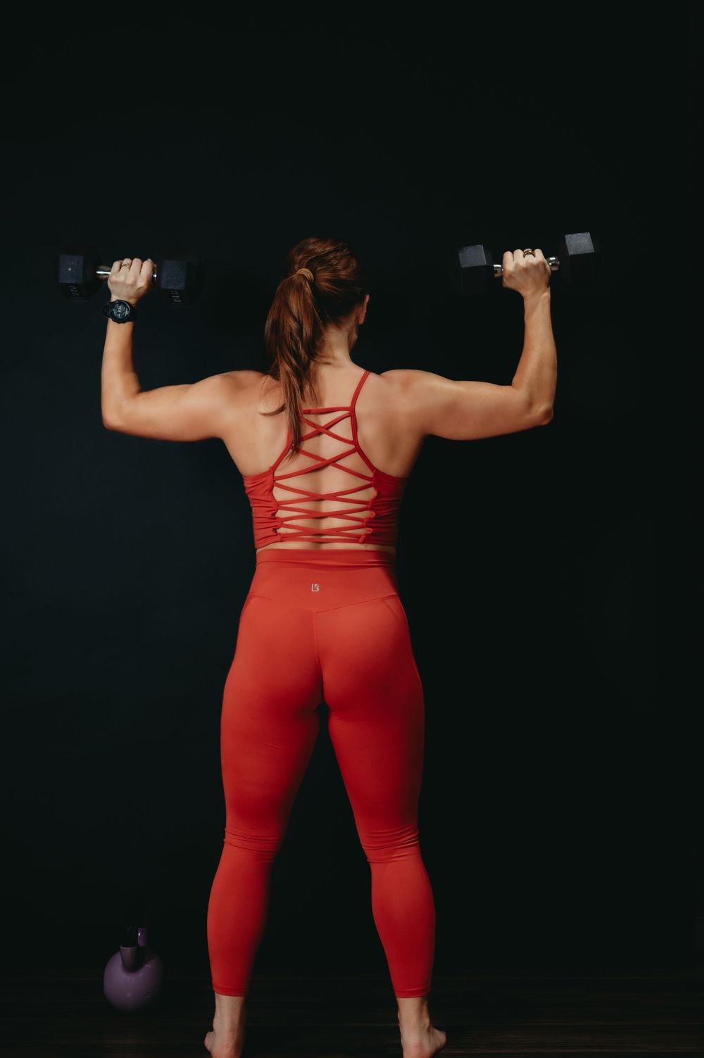 Person exercising with dumbbells wearing red strappy workout outfit against black background.