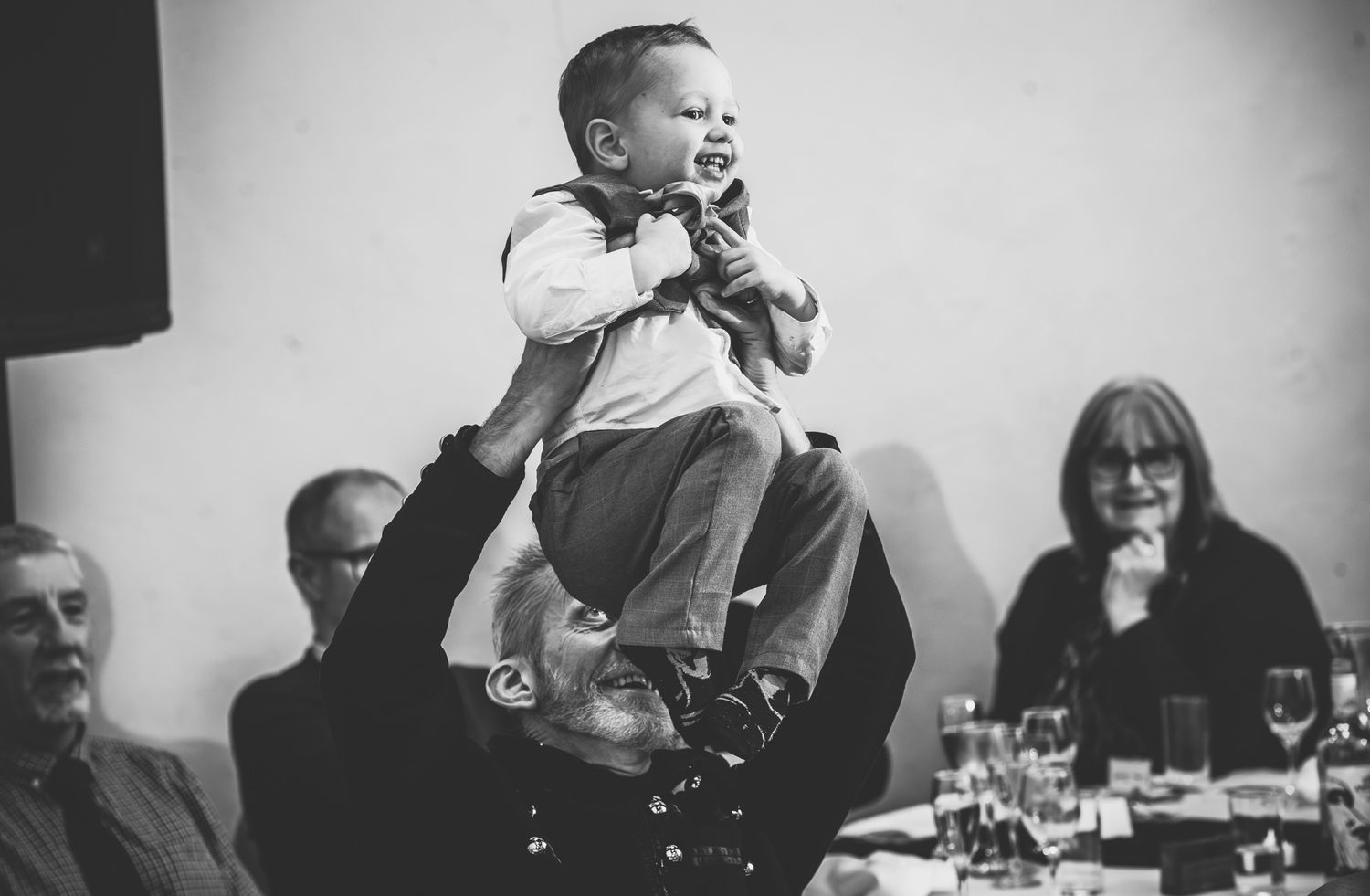 A joyful baby is lifted high in the air during what appears to be a celebratory dinner gathering.