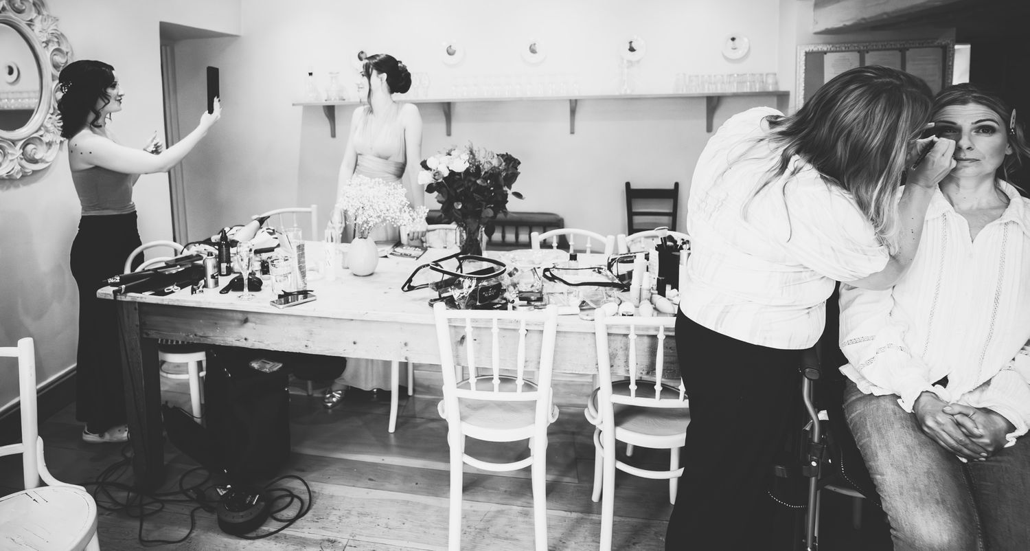 A busy backstage dressing room scene with people getting ready, surrounded by makeup tables and white chairs.