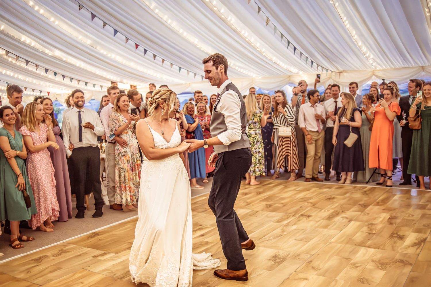 Wedding guests dance together in a large white tent reception venue with wooden flooring.