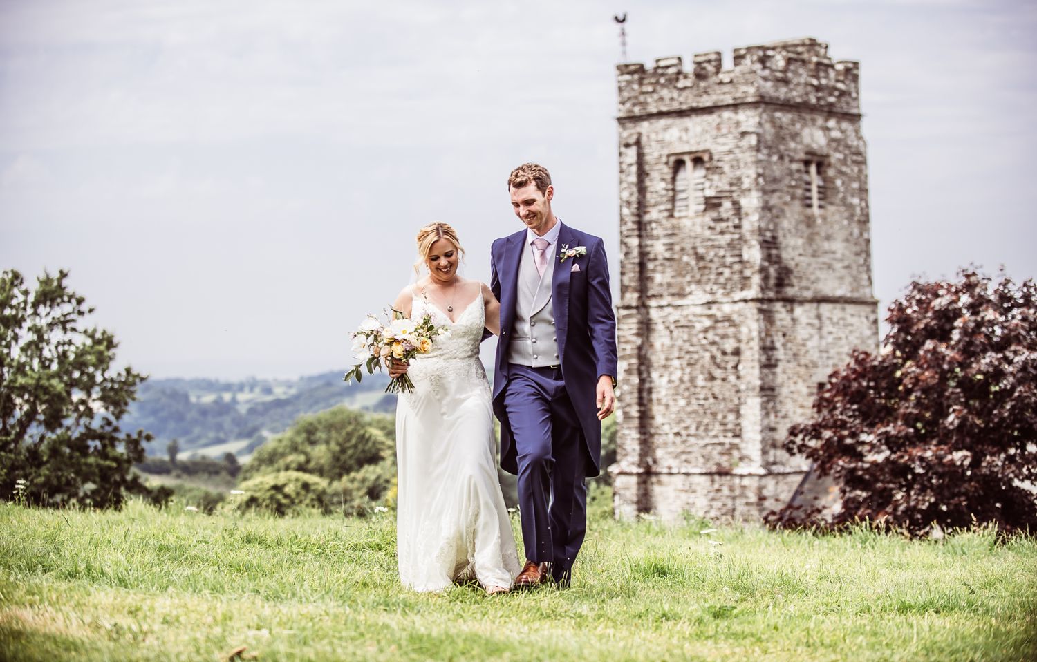 A couple celebrates their wedding day in front of a historic stone church tower in the countryside.