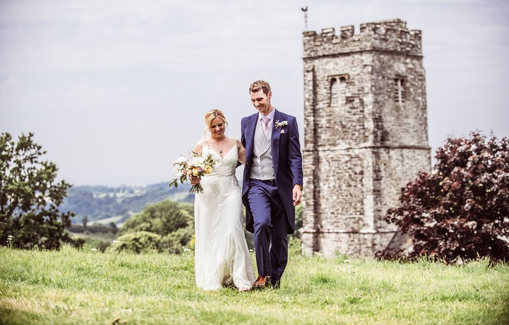 A couple celebrates their wedding day in front of a historic stone church tower in the countryside.
