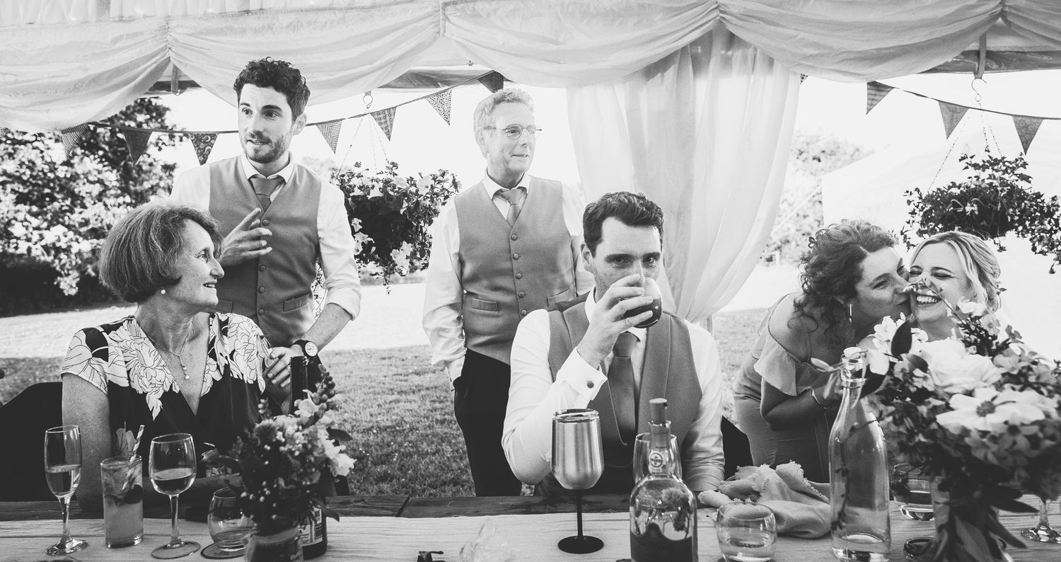 A black and white photograph of guests enjoying drinks at an outdoor wedding reception under a tent.