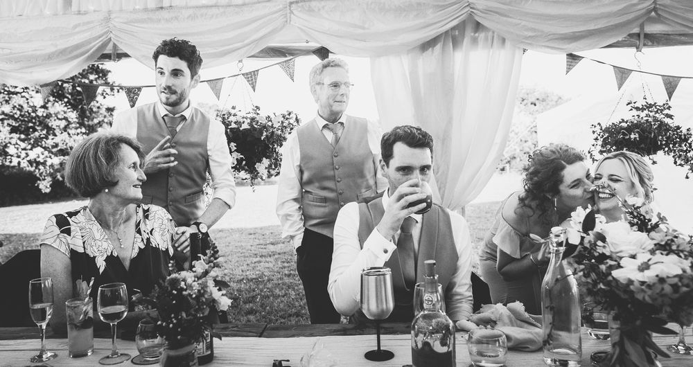 A black and white photograph of guests enjoying drinks at an outdoor wedding reception under a tent.