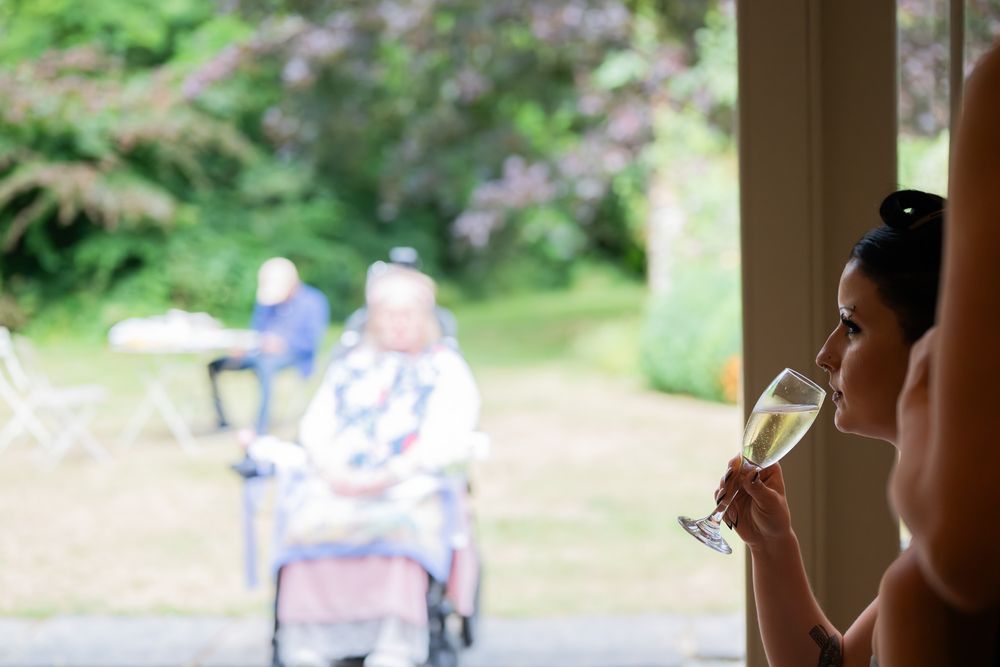 Person sips champagne while looking through window at blurred figures in garden setting.