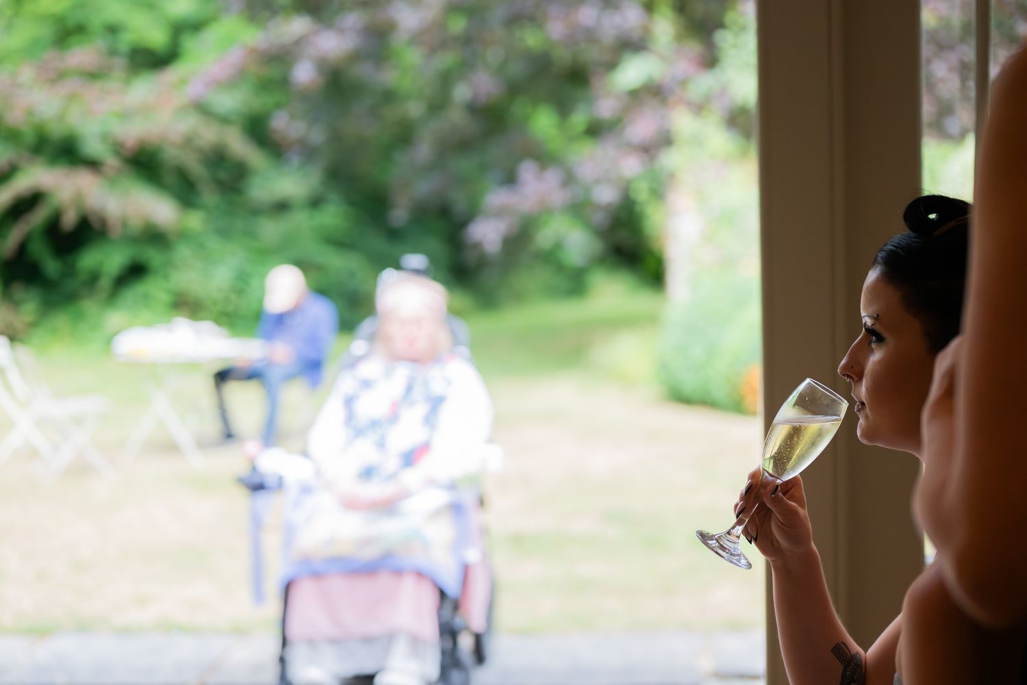 Person sips champagne while looking through window at blurred figures in garden setting.