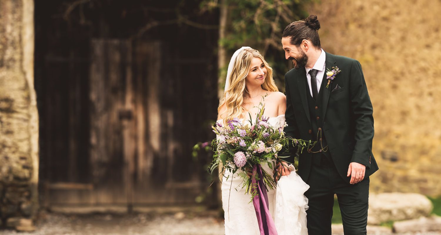 A couple in wedding attire walks together along a rustic stone wall during their outdoor ceremony.