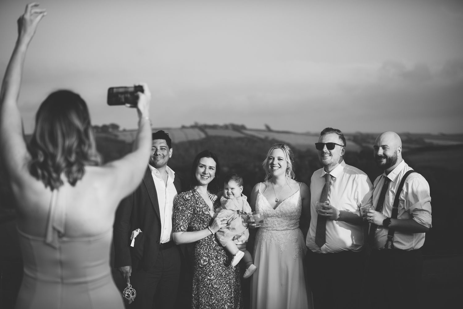 Black and white wedding celebration scene with guests mingling outdoors against a scenic backdrop.