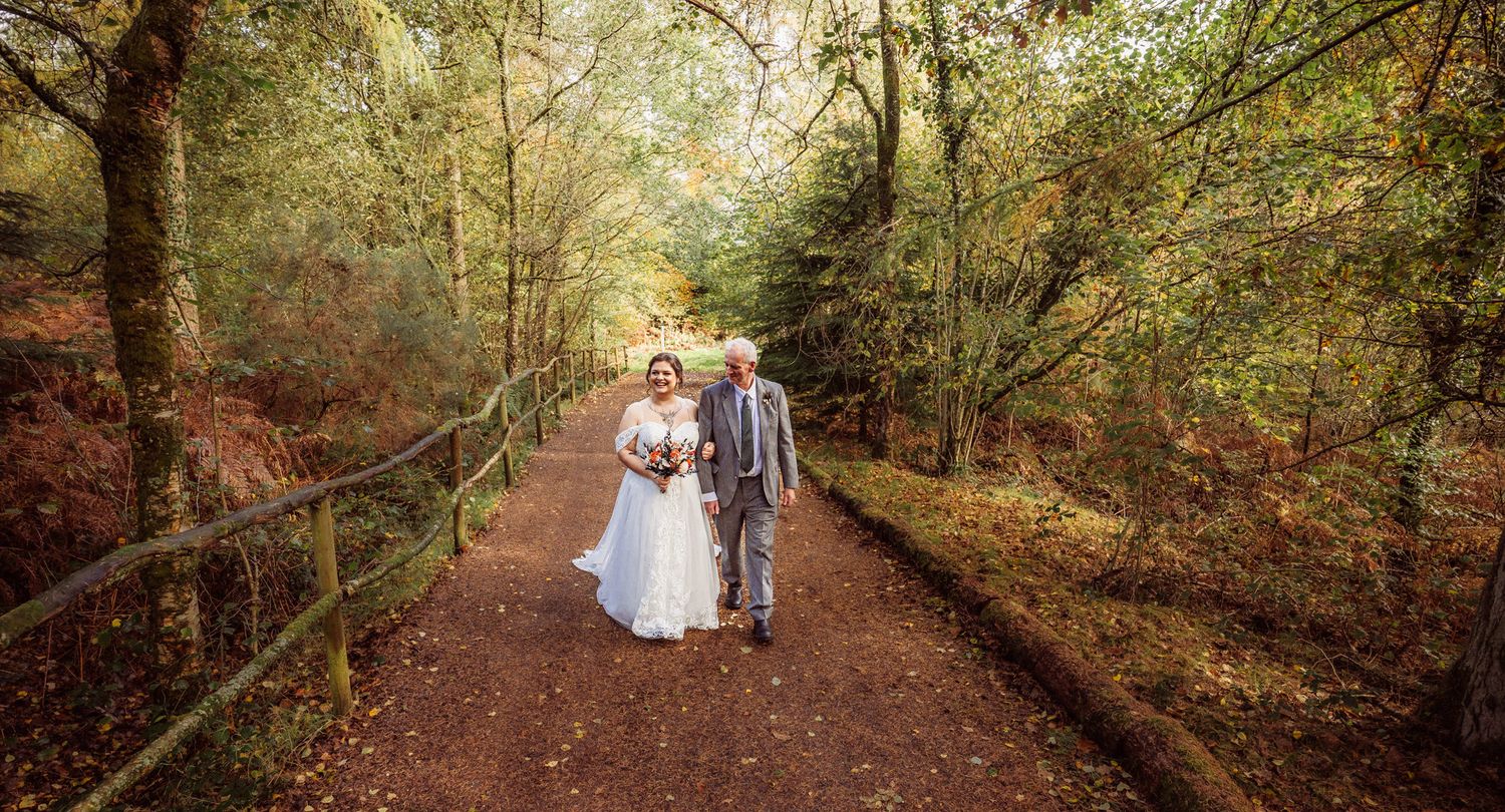 A couple walks hand in hand down a wooded autumn path surrounded by golden foliage and natural light.