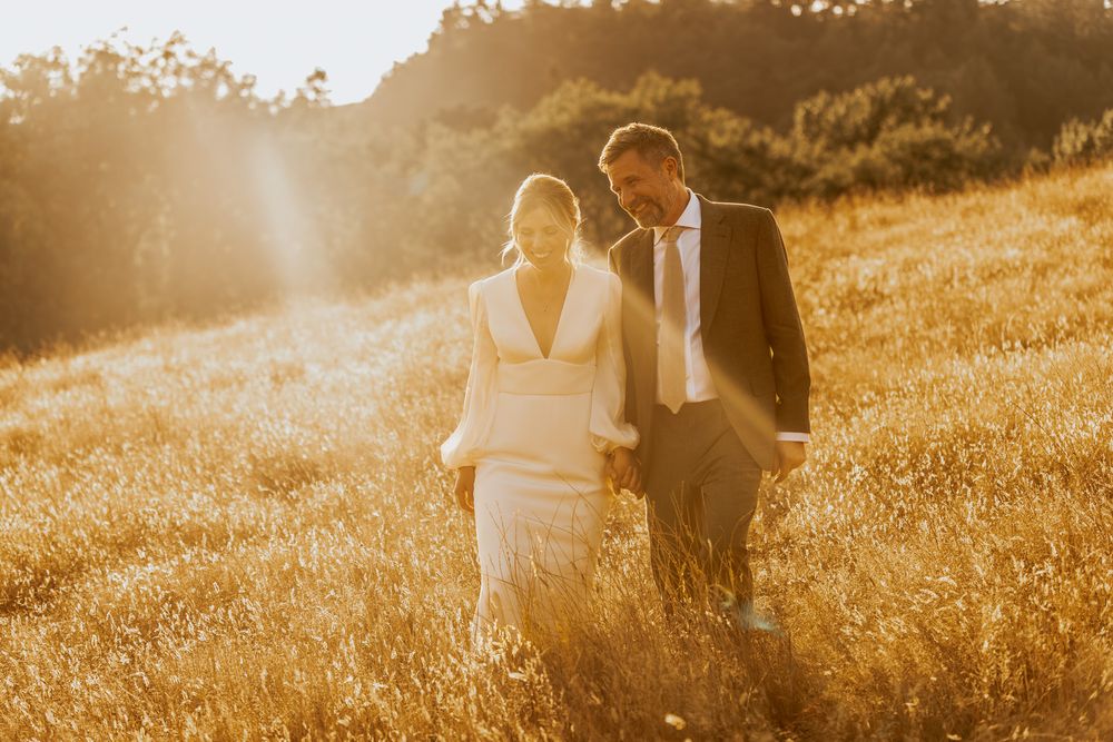 A couple walks through a golden sunlit meadow during sunset in formal wedding attire.
