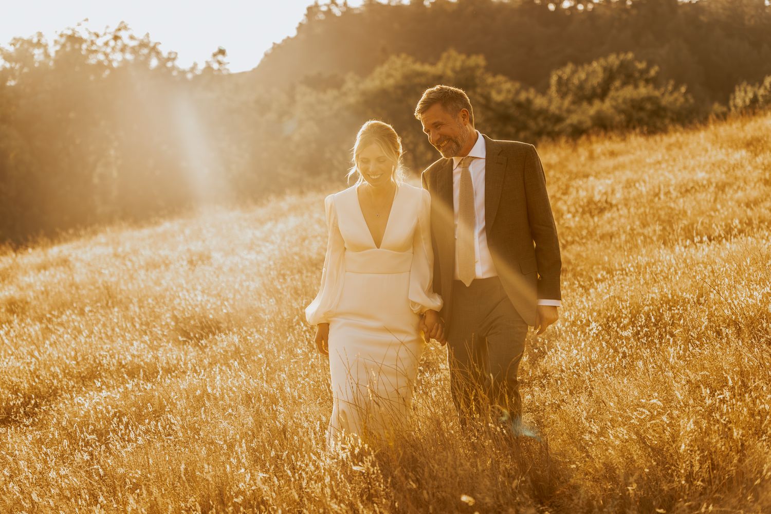 A couple walks through a golden sunlit meadow during sunset in formal wedding attire.