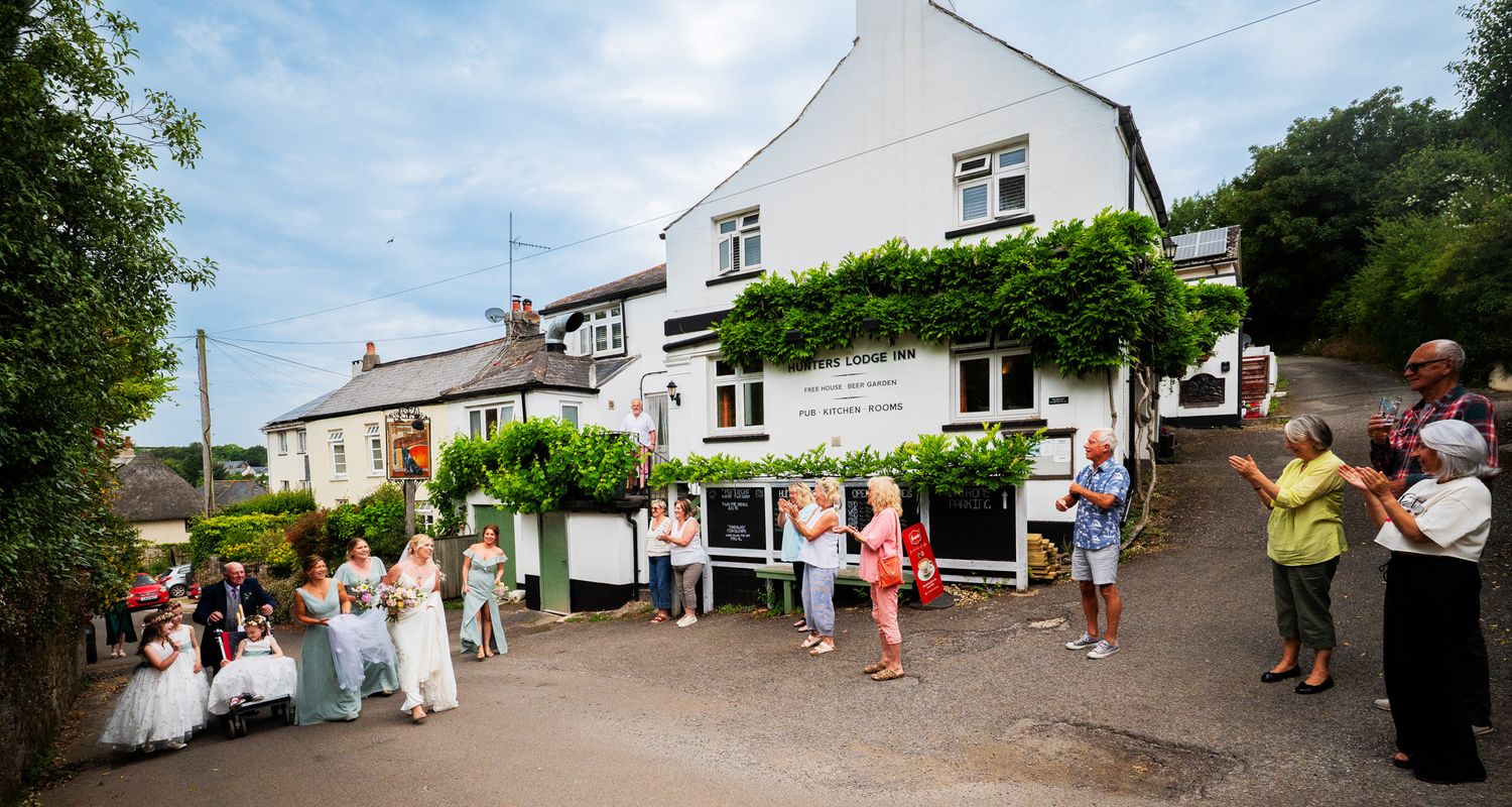 Wedding guests mingle and socialize on a charming village street lined with white buildings and trees.