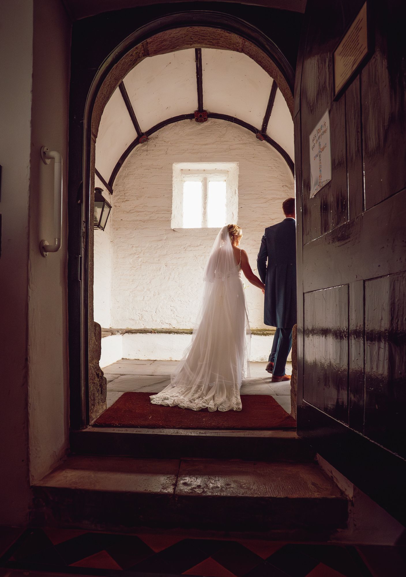 A couple in wedding attire stand in an arched doorway with white walls and red carpet, captured in romantic lighting.