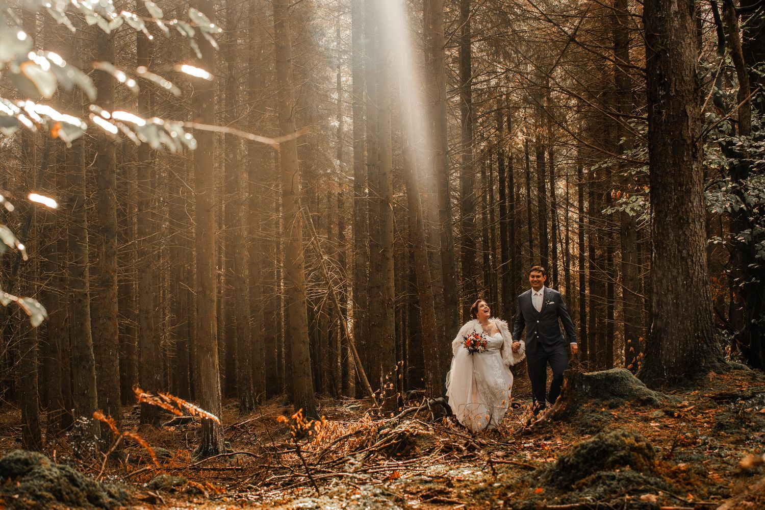 A couple walks through a misty autumn forest with sunlight streaming through tall trees in warm golden tones.