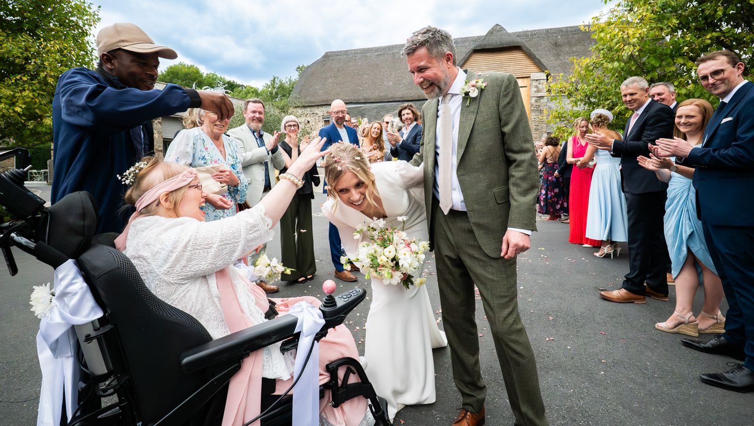 Wedding guests throw confetti at a couple during an outdoor celebration on a sunny day.
