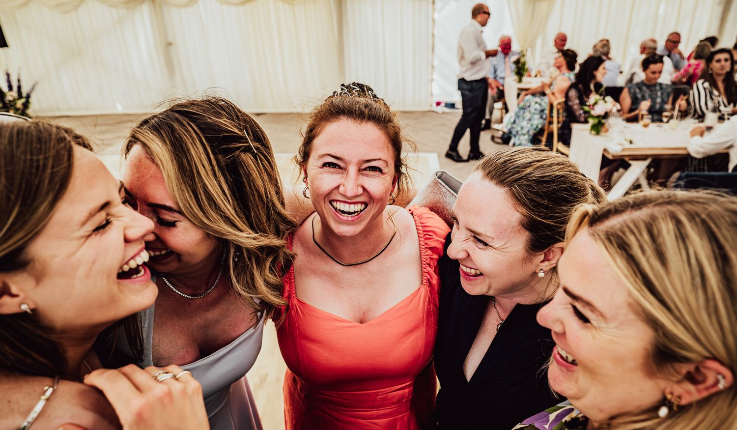 A group of friends laugh together joyfully at a wedding reception in a white tent venue.