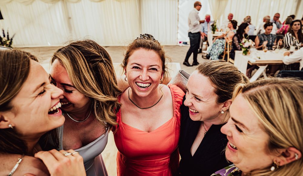 A group of friends laugh together joyfully at a wedding reception in a white tent venue.